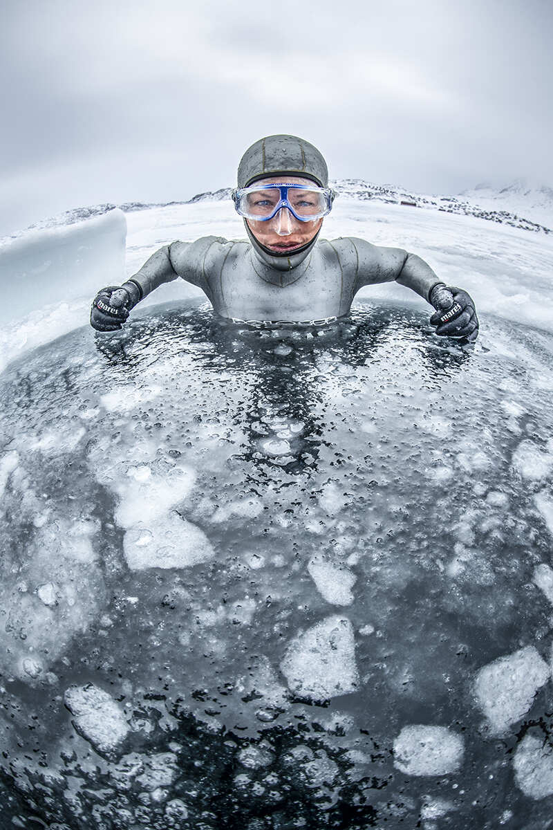A diver wearing goggles and a wetsuit sits half submerged in ice
